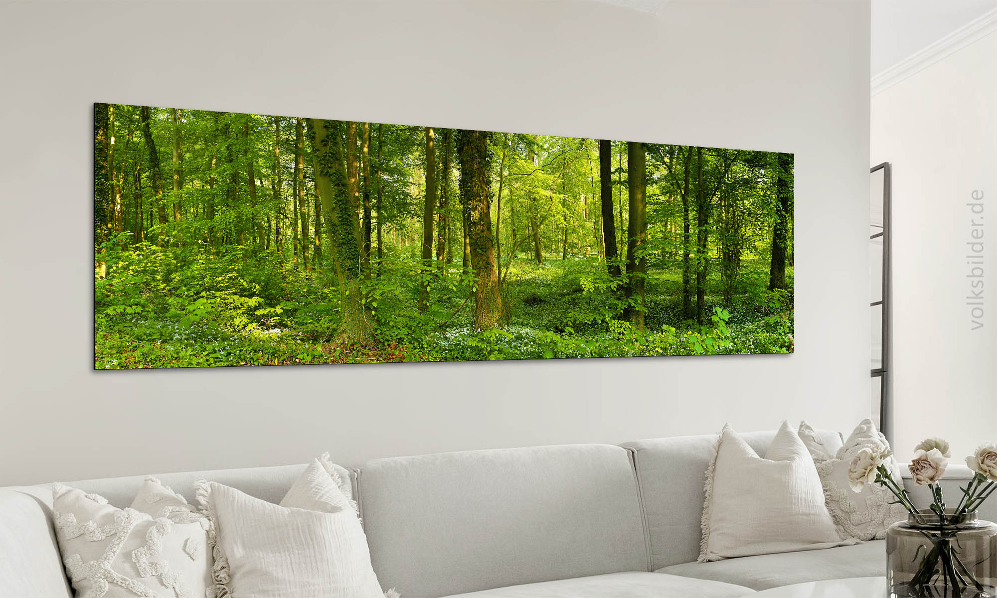 Foto eines Wohnzimmers. An der Wand steht eine Couch mit hellgrau-beigem Stoffbezug und mehreren Kissen in derselben Farbe. Über der Couch hängt ein langes, großes Panorama-Wandbild. Das Motiv des Wandbildes ist ein Auwald im Frühling.