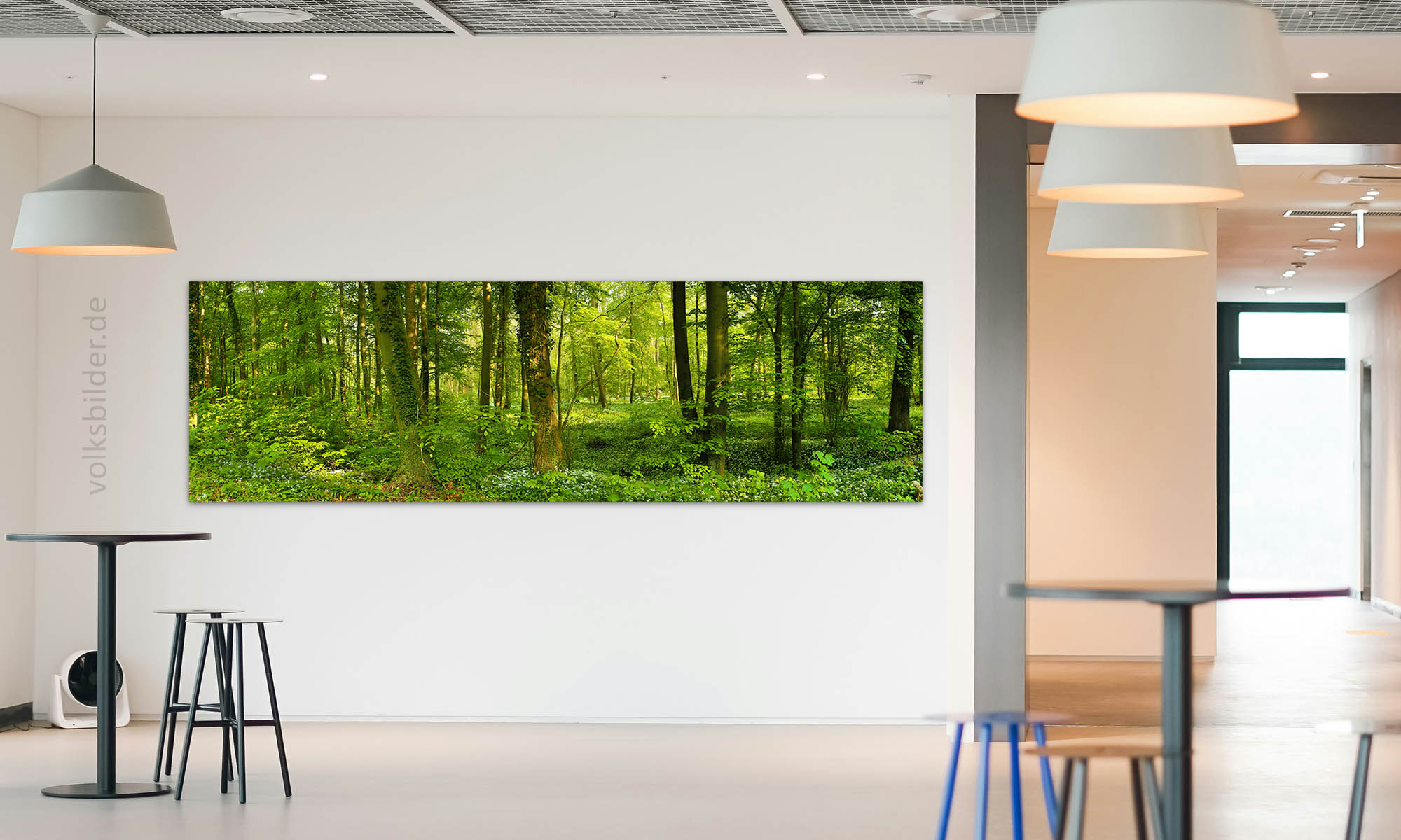 Foto eines großen Aufenthaltsraumes in einer Firma. An der Decke hängen grpße weiße Lampen, links und rechts im Bild seiht man Tische mit Barhockern. An der Wand im Hintergrund hängt ein langes, großes Panorama-Wandbild. Das Motiv des Wandbildes ist ein Auwald im Frühling.