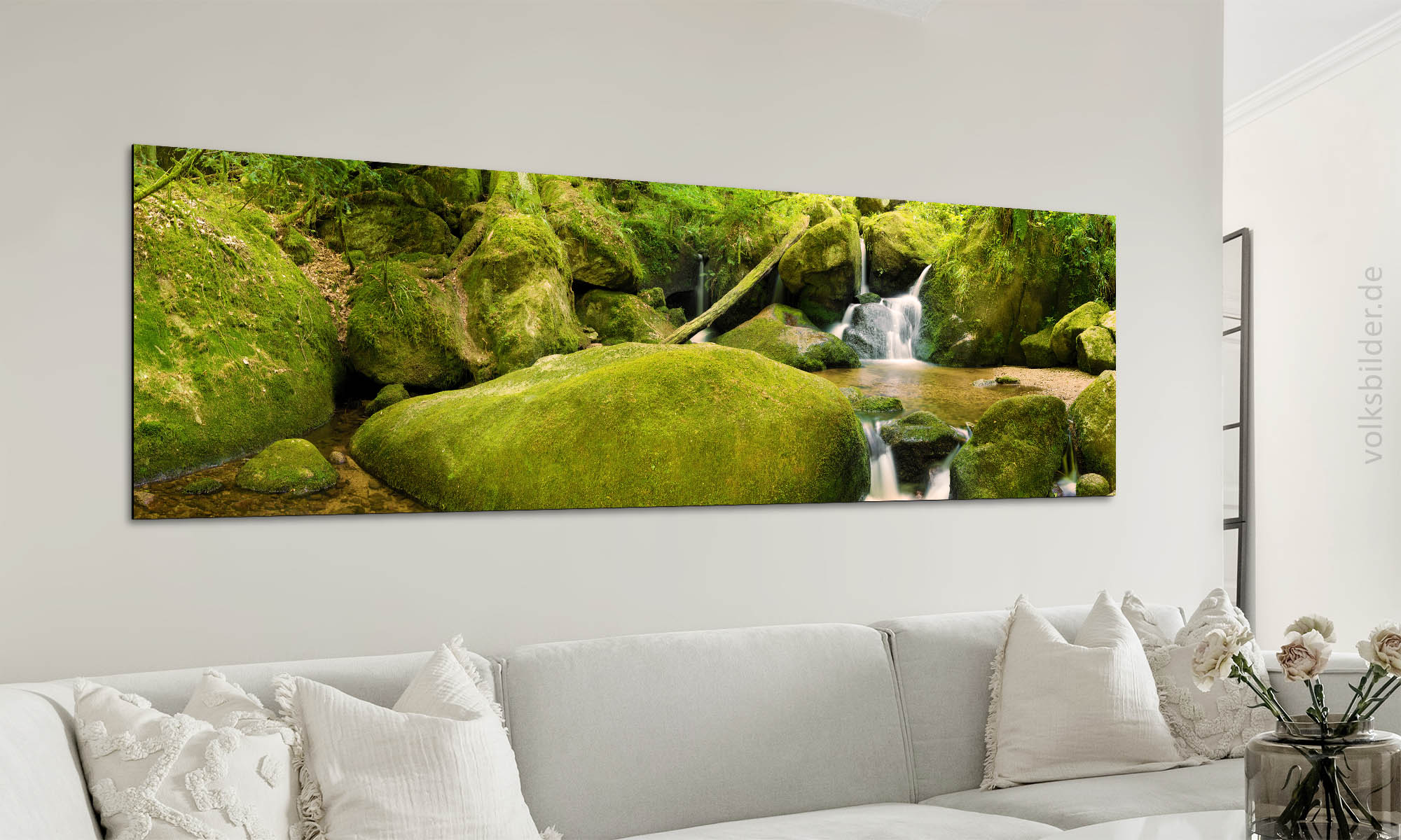 Foto von einem Wohnzimmer. Im Hintergrund hängt an der Wand über einer Couch mit Kissen ein langes, breites Wandbild. Das Wandbild zeigt einen Bach in felsiger Umgebung in einem Wald.