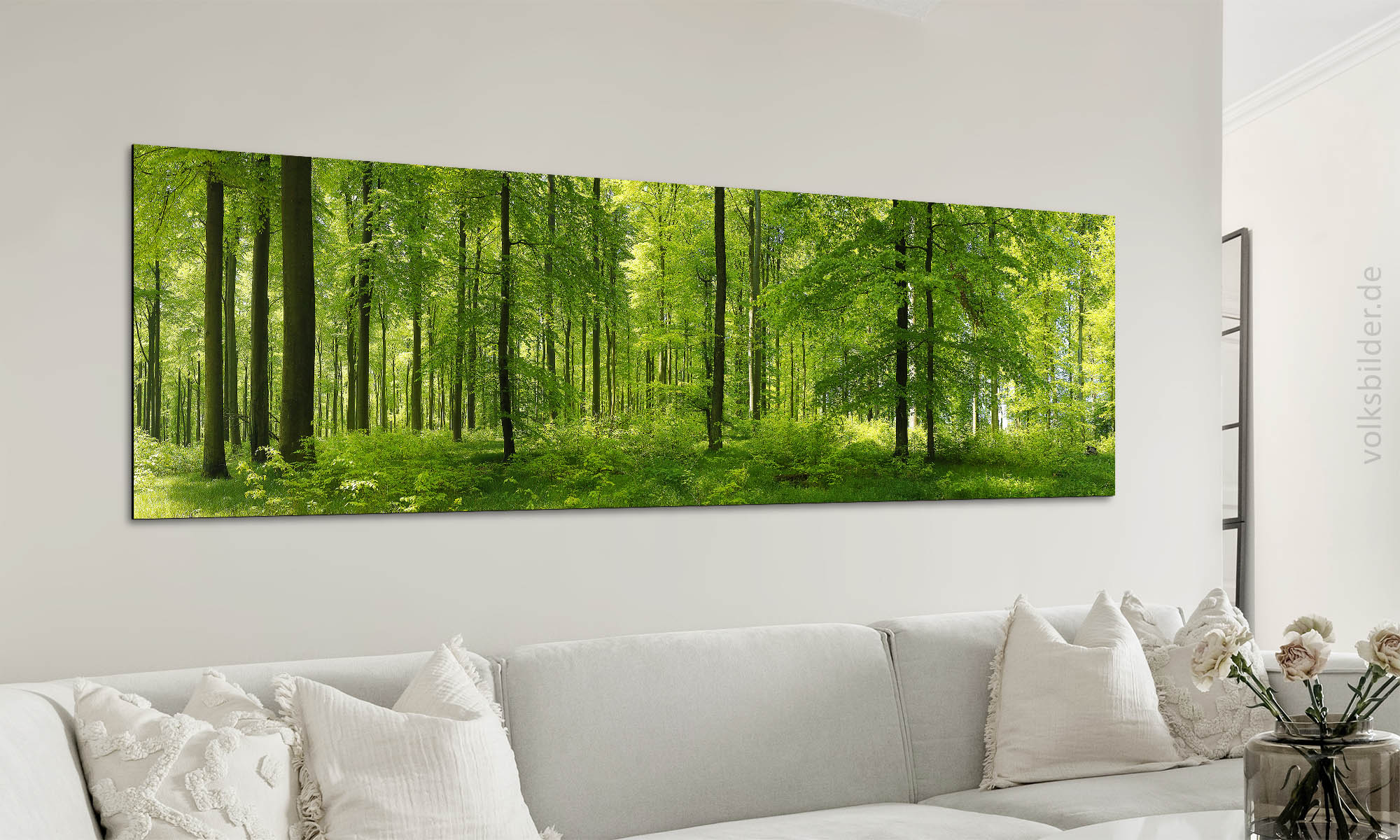 Das Foto zeigt ein Wohnzimmer. An der Wand steht eine Couch mit vielen Kissen. An der Wand über der Couch hängt ein langes, großes Panorama-Wandbild. Es zeigt einen Buchenwald im Frühjahr mit alten und jungen Bäumen. Das Laub der Bäume ist leuchtend hellgrün. Der Waldboden ist mit Seegras bedeckt.