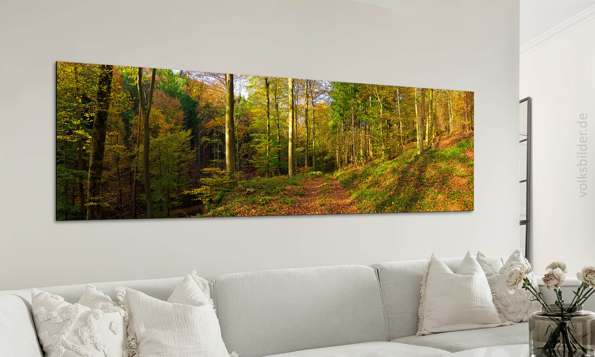 Foto eines Wohnzimmers. An der Wand steht eine hell-beige Couch mit mehreren Kissen. Darüber hängt ein langes, großes Panorama-Wandbild. Es zeigt den herbstlich bunten Wald bei Edenkoben in Rheinland-Pfalz.