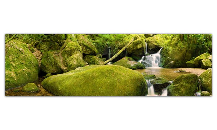 Ein längliches, großes Panoramabild. Auf dem Bild ist ein kleiner Bach zu sehen, der im Wald durch Steine und Felsen fließt.   